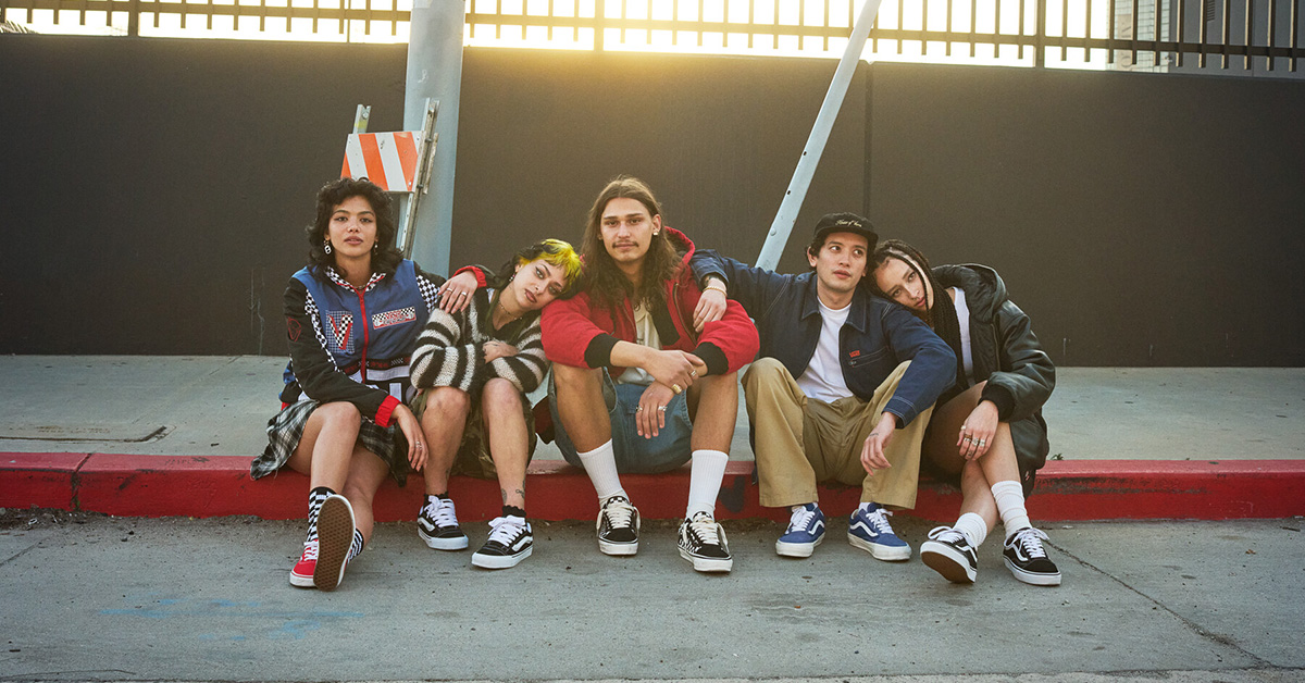 Grupa pięciu młodych osób w casualowych stylizacjach streetwear siedzi razem na krawężniku miejskiego chodnika.