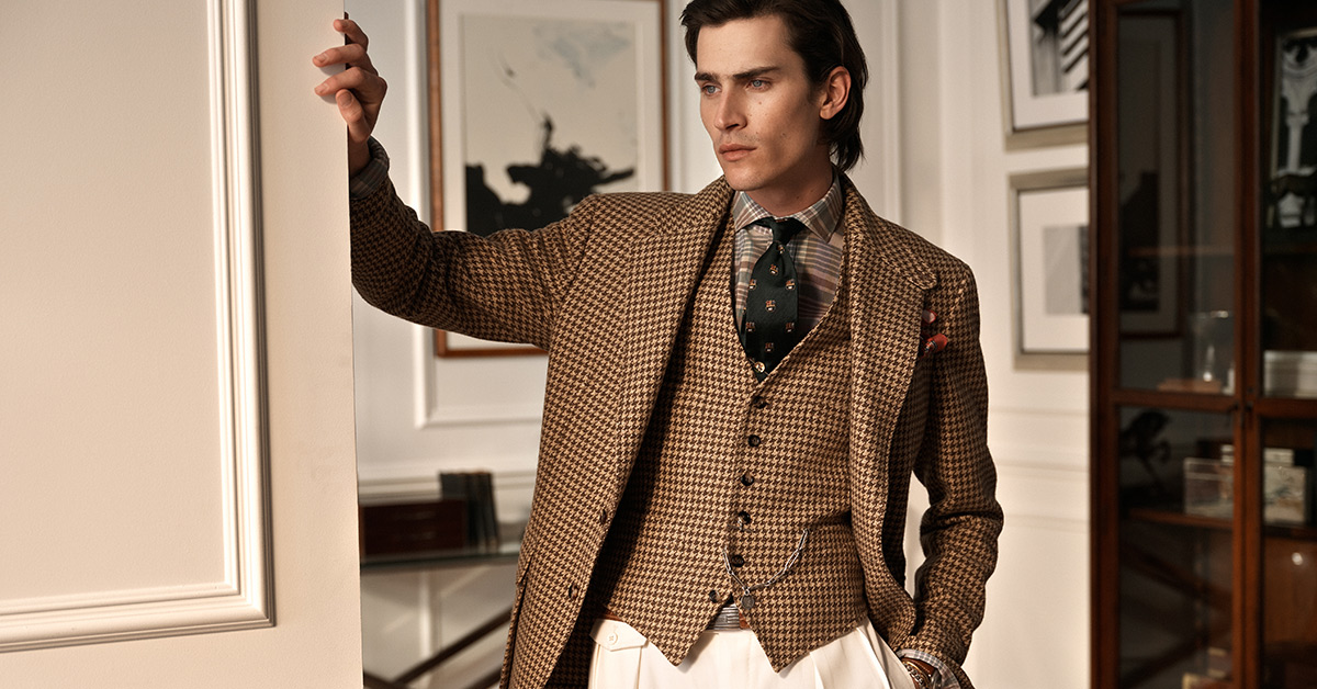 Elegancko ubrany mężczyzna w stylowym garniturze w pepitkę, z klasyczną kamizelką i białymi spodniami, pozuje w luksusowym wnętrzu gabinetu.