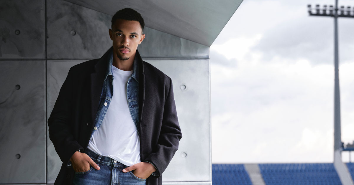 Mężczyzna w stylizacji casual – ciemny płaszcz, jeansowa kurtka warstwowo na białym t-shircie i niebieskie jeansy, miejski plener ze stadionem w tle, minimalistyczna męska moda jesień/zima