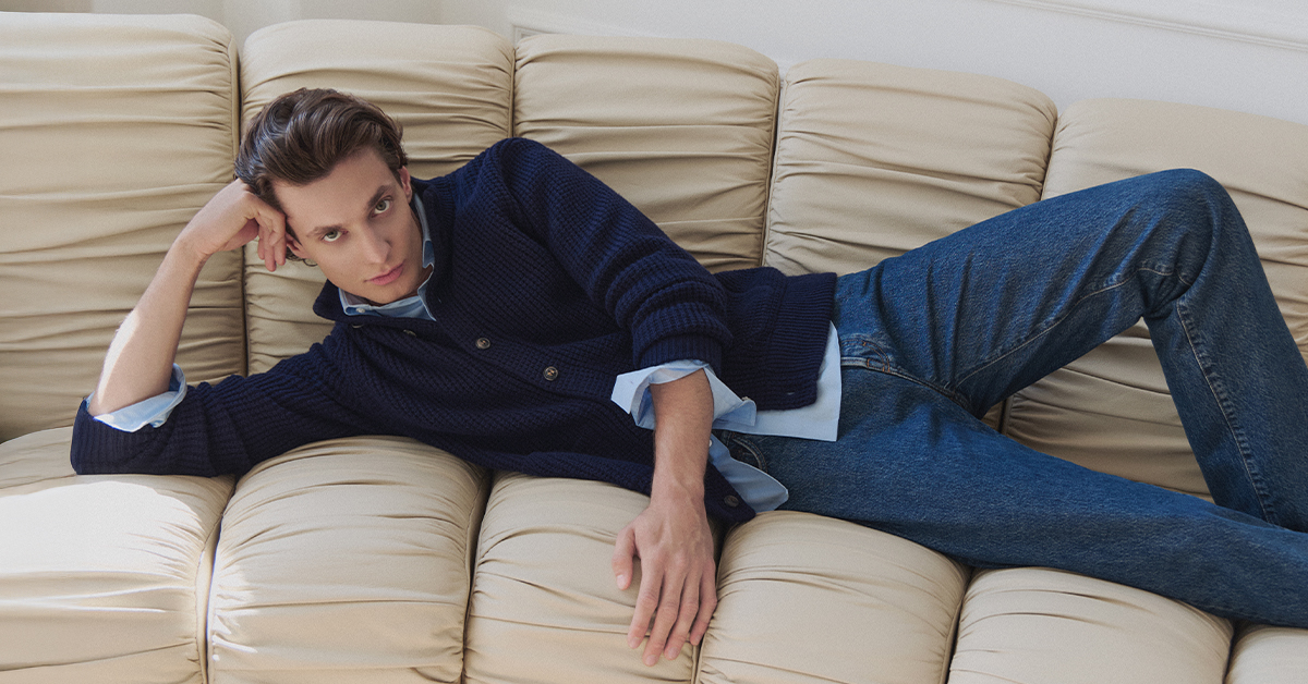 Mężczyzna w granatowym kardiganie i jasnoniebieskiej koszuli, denimowe jeansy, leżący na beżowej sofie, casualowa stylizacja męska, minimalistyczna moda sezonu jesień