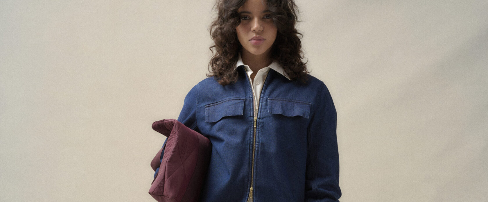 Młoda kobieta w komplecie Barbour: granatowa denimowa overshirt Arla z suwakiem, biała koszula Evette, szerokie jeansowe spodnie Arla i bordowa pikowana torba Healy, minimalistyczna stylizacja na jednolitym tle.
