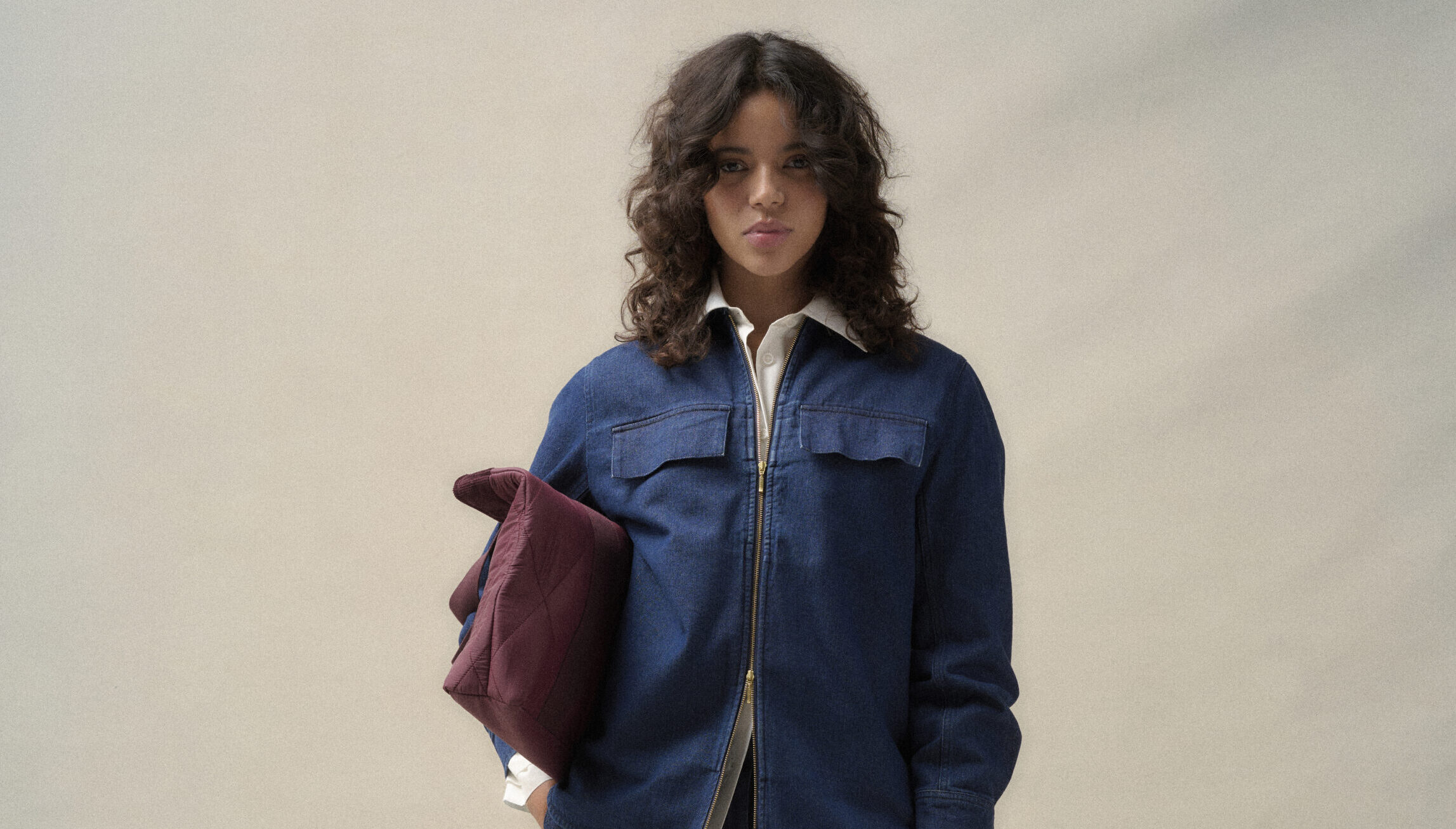 Młoda kobieta w komplecie Barbour: granatowa denimowa overshirt Arla z suwakiem, biała koszula Evette, szerokie jeansowe spodnie Arla i bordowa pikowana torba Healy, minimalistyczna stylizacja na jednolitym tle.