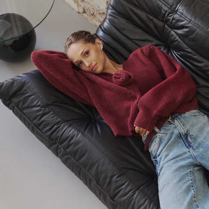 Kobieta w bordowym wełnianym kardiganie z kwiatową aplikacją, niebieskie mom jeans, leży na czarnej skórzanej sofie; biżuteria srebrna, jesienna stylizacja casual, nowoczesne wnętrze