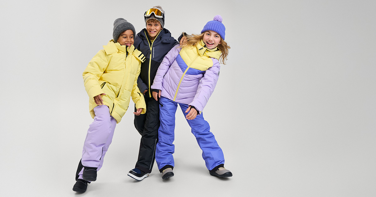 Troje dzieci w jasnych, pastelowych zimowych kombinezonach, kurtkach i czapkach; dziewczynka w fioletowo-żółtym zestawie, chłopiec w ciemnej kurtce i goglach, chłopiec w żółtej kurtce i szarej czapce, radosna poza – dziecięca moda zimowa, kolekcja narciarska