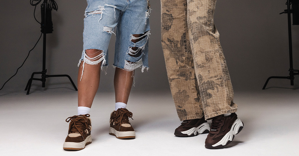 Dwie osoby w modnych sneakersach – jedna w brązowych butach do biegania, szerokich beżowych spodniach z motywem florystycznym; druga w brązowych sneakersach, białych skarpetkach i mocno podartych denimowych bermudach; streetwearowa stylizacja unisex