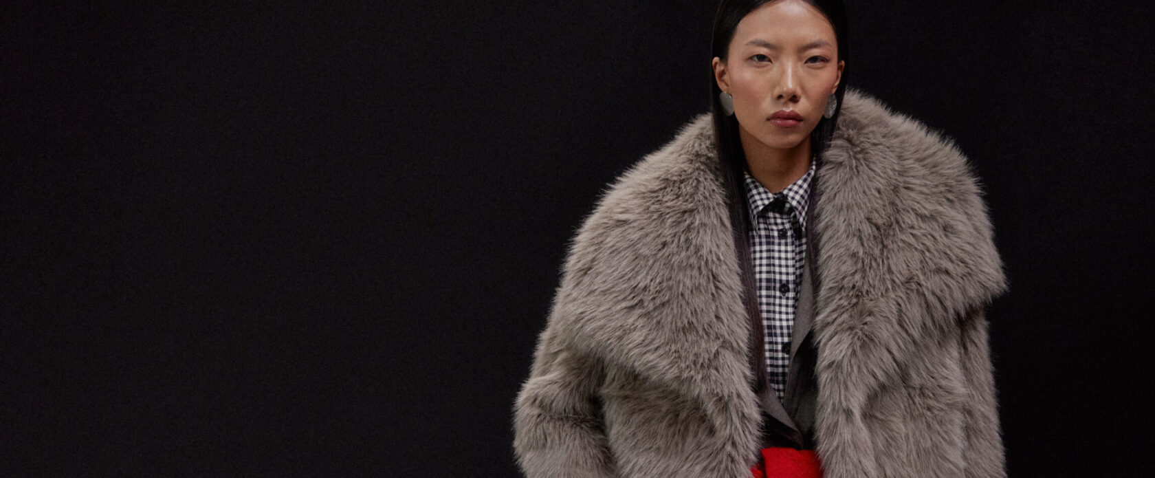 Kobieta w obszernej szarej futrzanej kurtce na czarnym tle, koszula w kratę, czerwony akcent w talii, czarne spodnie z szeroką nogawką, jasnobeżowa torebka na ramię – stylizacja wieczorowa jesień/zima, elegancja, warstwy, faux fur trend