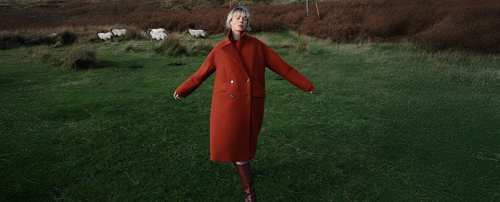 Kobieta w długim rudym płaszczu oversize, brązowe kozaki, jesienny krajobraz, owce na pastwisku w tle – minimalistyczna stylizacja na jesień/zima, naturalne kolory, country chic