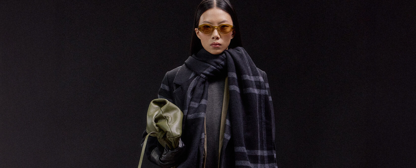 Kobieta w ciemnym wełnianym płaszczu, szeroki szal w kratę, szara dzianina, trzyma oliwkową torebkę i czarne rękawiczki – minimalistyczna stylizacja jesień/zima, odcienie khaki, trendy warstwowe, tło czarne