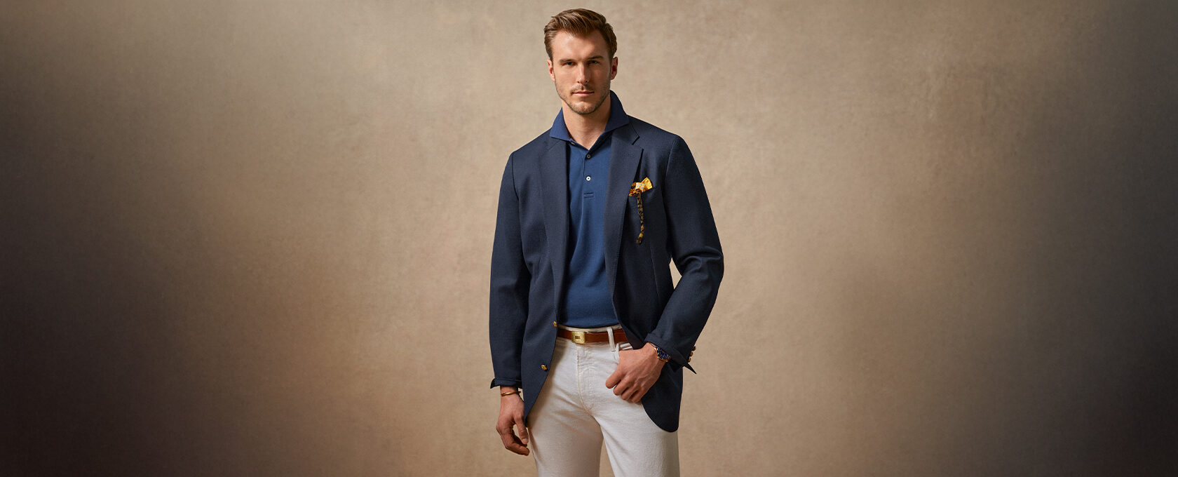 Mężczyzna w granatowej marynarce, granatowa koszulka polo, białe spodnie, brązowy pasek; elegancki smart casual, żółta poszetka – moda męska na wiosnę/lato 2025, outfit do pracy i spotkań