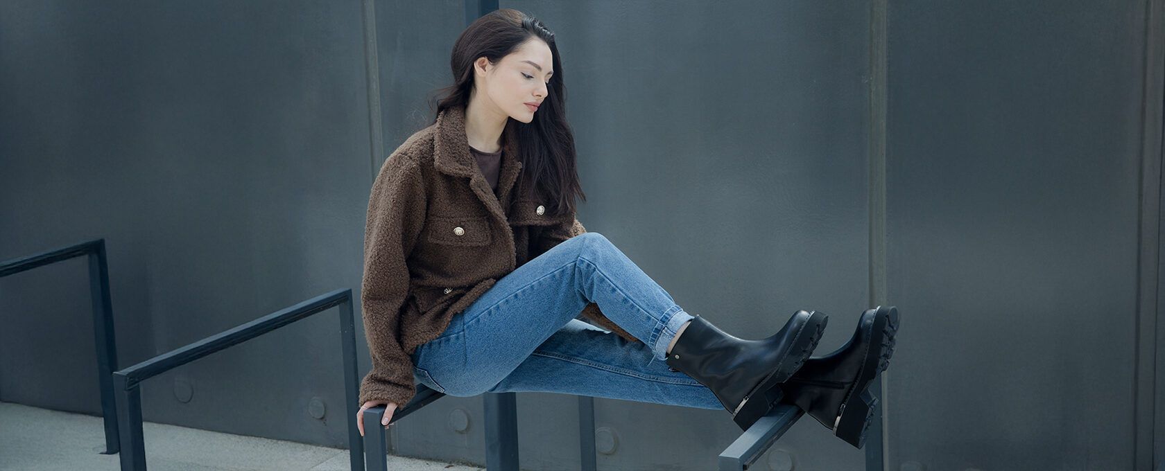 Kobieta w brązowej kurtce o futrzanej fakturze, niebieskie jeansy, czarne masywne kozaki, minimalistyczna stylizacja na tle szarej ściany – moda casual jesień/zima