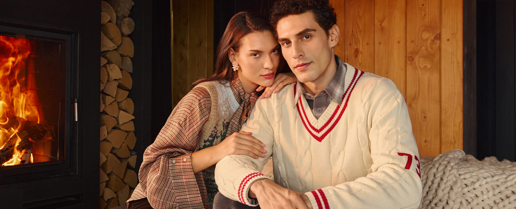 Dwoje młodych ludzi w stylowych ubraniach siedzi przy kominku – kobieta w kratkowanej bluzce i wzorzystej narzutce, mężczyzna w kremowym swetrze z bordowym detalem i koszuli, klimat hygge, jesienne wieczory