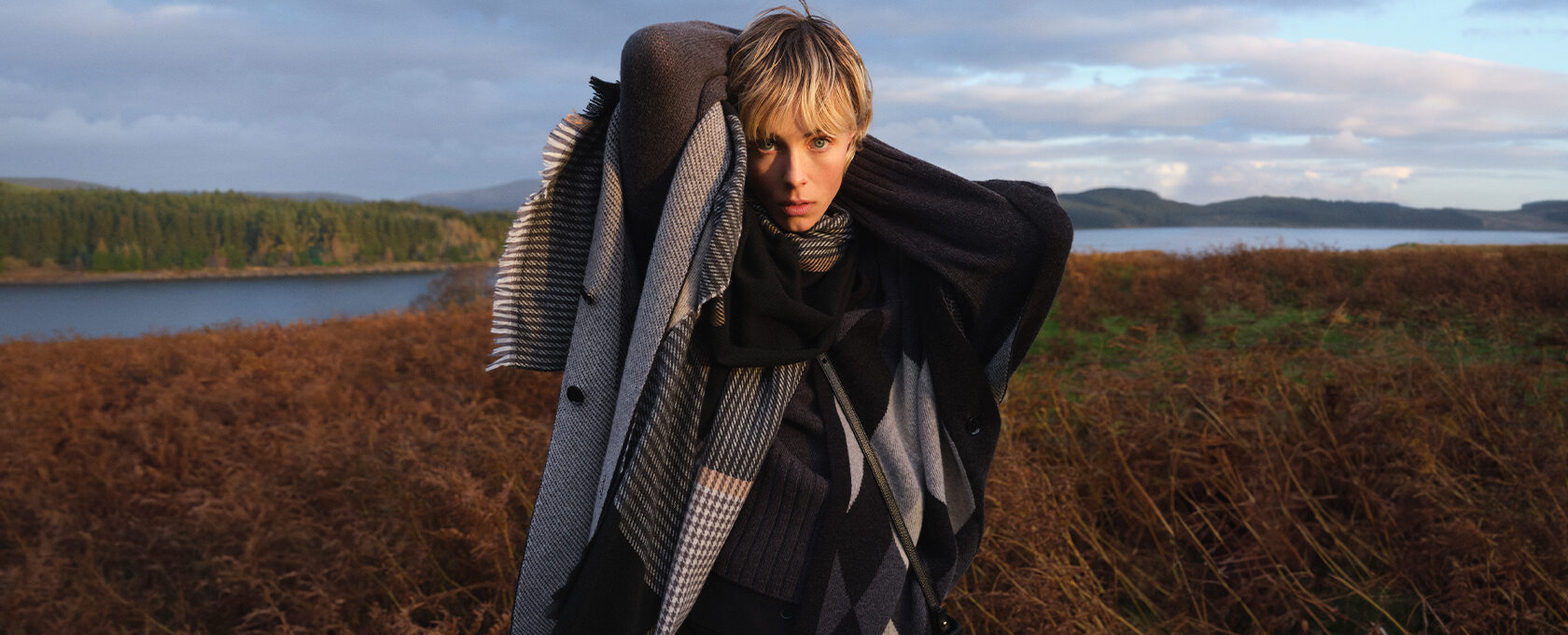 Osoba owinięta w ciepłe, warstwowe szale w odcieniach szarości i czerni, oversize płaszcz, naturalny krajobraz: jezioro, brązowe trawy i las; stylizacja jesienno-zimowa, moda outdoor, hygge